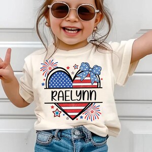 May include: A cream-colored t-shirt featuring a patriotic heart design with the name "RAELYNN" in black. The heart is divided into sections with the American flag and a blue bow. Fireworks and stars surround the heart.