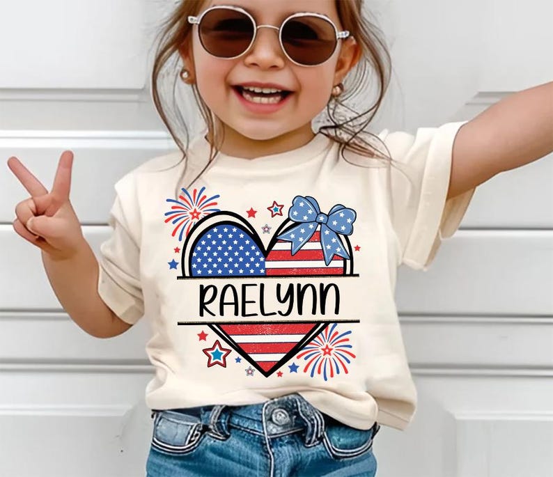 May include: A cream-colored t-shirt featuring a patriotic heart design with the name "RAELYNN" in black. The heart is divided into sections with the American flag and a blue bow. Fireworks and stars surround the heart.