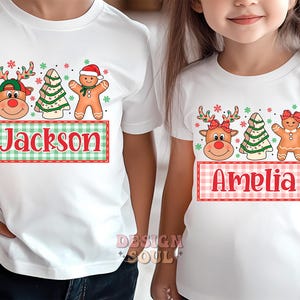 May include: Two white t-shirts with a Christmas theme. One shirt features a reindeer, a gingerbread person, and a Christmas tree with the name "Jackson" on a plaid banner. The other shirt features a reindeer, a gingerbread person, and a Christmas tree with the name "Amelia" on a plaid banner.