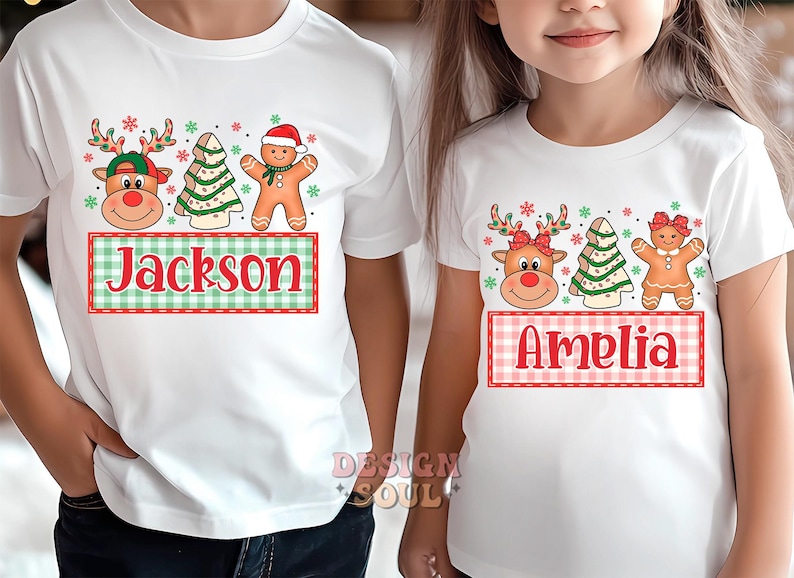 May include: Two white t-shirts with a Christmas theme. One shirt features a reindeer, a gingerbread person, and a Christmas tree with the name "Jackson" on a plaid banner. The other shirt features a reindeer, a gingerbread person, and a Christmas tree with the name "Amelia" on a plaid banner.