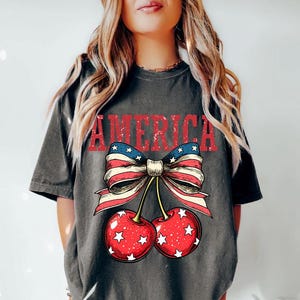 May include: A gray t-shirt with a distressed graphic of a red, white, and blue bow with cherries. The bow is made of an American flag pattern and the cherries have white stars on them. The word "AMERICA" is printed above the bow in red.