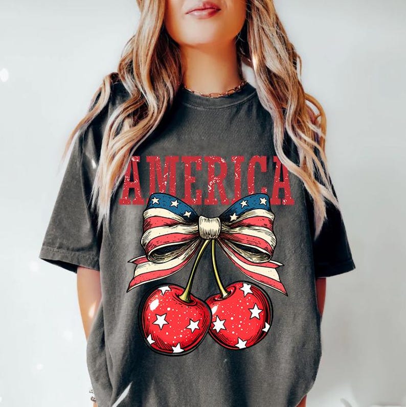 May include: A gray t-shirt with a distressed graphic of a red, white, and blue bow with cherries. The bow is made of an American flag pattern and the cherries have white stars on them. The word "AMERICA" is printed above the bow in red.