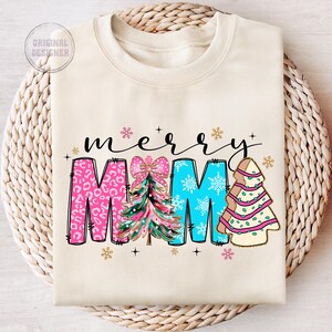 Peut inclure: Un sweat-shirt cr&egrave;me avec le texte "merry mama" en lettres noires. Les lettres sont d&eacute;cor&eacute;es de sapins de No&euml;l roses, bleus et verts.