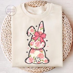 May include: A cream-colored sweatshirt with a floral bunny design. The bunny is pink and white with a pink bow. The bunny is surrounded by pink flowers.