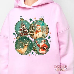 May include: Pink hoodie featuring a Christmas design with four ornaments depicting a snowman, reindeer, Santa Claus, and a Christmas tree. The ornaments are surrounded by stars.