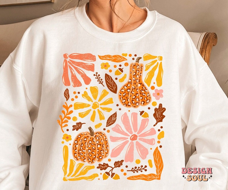 May include: White sweatshirt with a graphic print of a square filled with orange, yellow, pink, and brown floral and pumpkin designs. The text "DESIGN SOUL" is printed in the bottom right corner.