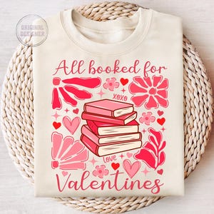 May include: A cream-colored sweatshirt with the text "All booked for Valentines" in pink lettering. The design features a stack of pink books with the words "XOXO" and "Love" on them, surrounded by pink flowers and hearts.
