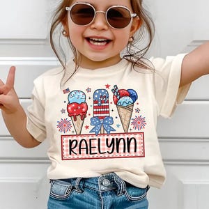 May include: A white t-shirt with a red, white, and blue ice cream cone design. The shirt has the name "Raelynn" written on it.