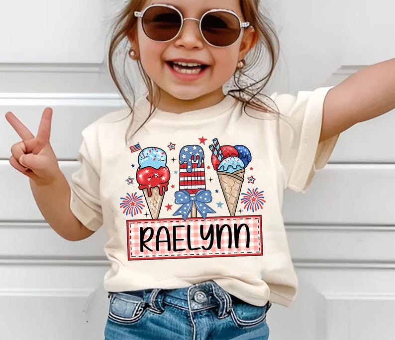 May include: A white t-shirt with a red, white, and blue ice cream cone design. The shirt has the name "Raelynn" written on it.