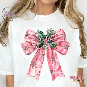 May include: A white t-shirt with a pink bow with holly and berries. The bow has a sparkly, watercolor effect.