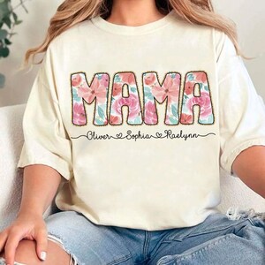 May include: A white t-shirt with the word "MAMA" in large pink letters with a floral pattern. The names "Oliver", "Sophia", and "Raelynn" are written below the word "MAMA" in a smaller font.