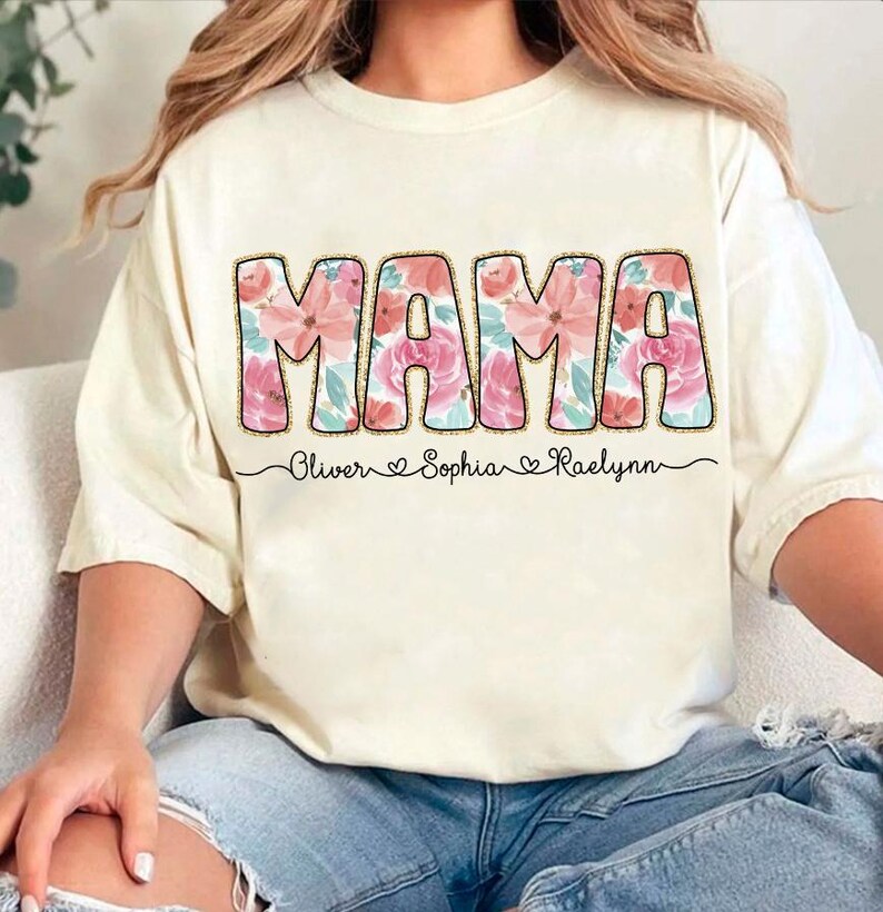 May include: A white t-shirt with the word "MAMA" in large pink letters with a floral pattern. The names "Oliver", "Sophia", and "Raelynn" are written below the word "MAMA" in a smaller font.