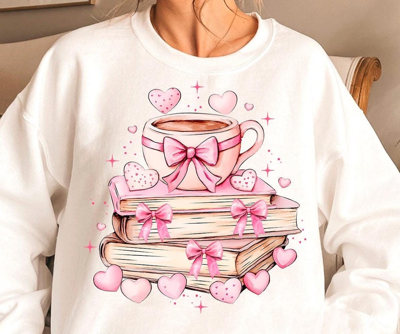 K&ouml;nnte beinhalten: Ein wei&szlig;er Pullover mit einer rosa und wei&szlig;en Illustration einer Tasse Kaffee, einem Stapel B&uuml;cher und Herzen.