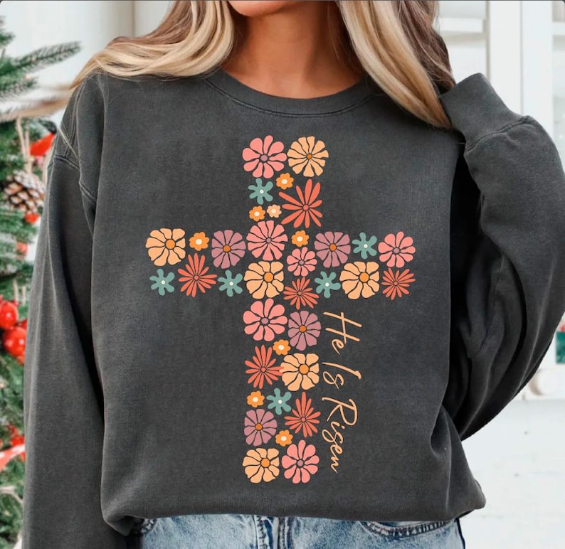 May include: A charcoal gray sweatshirt with a floral cross design and the text "He is Risen".