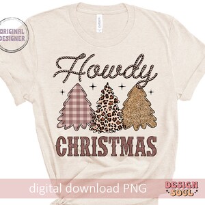 May include: A beige t-shirt with the text "Howdy Christmas" in pink and brown. The text is surrounded by three stylized Christmas trees in pink, brown, and gold.