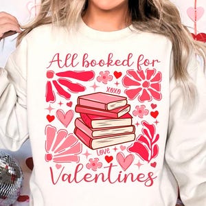 May include: White sweatshirt with a pink graphic design that says "All booked for Valentines". The graphic features a stack of books, hearts, flowers, and stars.