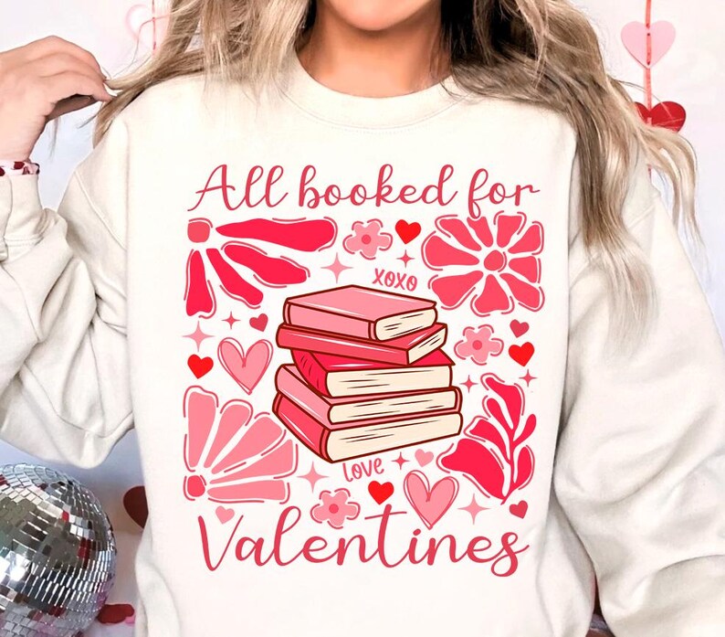 May include: White sweatshirt with a pink graphic design that says "All booked for Valentines". The graphic features a stack of books, hearts, flowers, and stars.