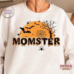 May include: White sweatshirt with a black and orange Halloween design. The design features a moon, bats, a spider web, a spider, and the text "MOMSTER".