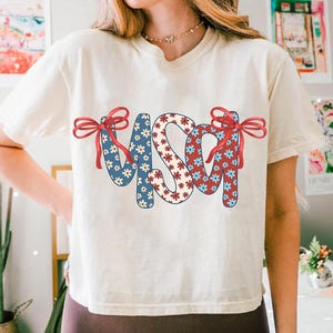May include: A white t-shirt with a red, white, and blue USA graphic. The letters are made of a floral pattern with red, white, and blue flowers. The graphic has red bows on each side.