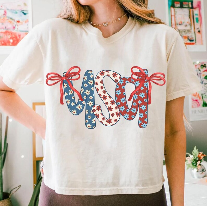 May include: A white t-shirt with a red, white, and blue USA graphic. The letters are made of a floral pattern with red, white, and blue flowers. The graphic has red bows on each side.