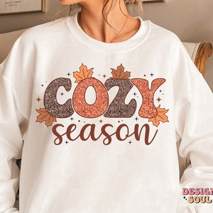 May include: White sweatshirt with the words "COZY season" in a fall-themed design. The text features glittery brown and orange letters, surrounded by autumn leaves and small star accents. A cozy fall-themed apparel item.