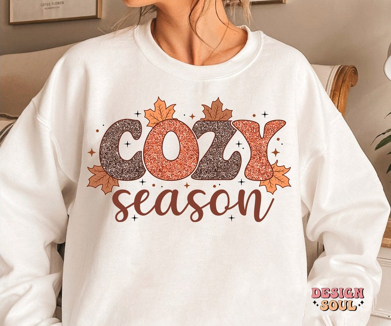 May include: White sweatshirt with the words "COZY season" in a fall-themed design. The text features glittery brown and orange letters, surrounded by autumn leaves and small star accents. A cozy fall-themed apparel item.