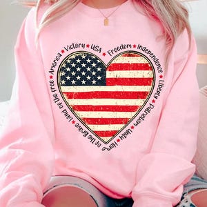 May include: Pink sweatshirt with a large heart-shaped American flag graphic. The heart is surrounded by the words "Home of the Brave, Land of the Free, America, Victory, USA, Freedom, Independence, Liberty, Patriotism, Unity".