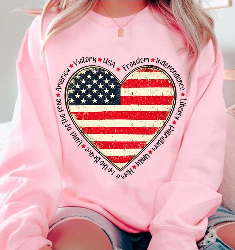 May include: Pink sweatshirt with a large heart-shaped American flag graphic. The heart is surrounded by the words "Home of the Brave, Land of the Free, America, Victory, USA, Freedom, Independence, Liberty, Patriotism, Unity".