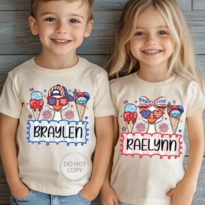 May include: Two children wearing beige t-shirts with patriotic designs. Each shirt features a cartoon duck wearing sunglasses and a hat or bow, with ice cream cones and the names "BRAYLEN" and "RAELYNN" in a decorative frame. The shirts are perfect for Independence Day.