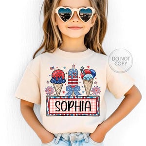 May include: A beige t-shirt featuring a patriotic ice cream design with red, white, and blue colors. The design includes ice cream cones, popsicles, and the name "SOPHIA" in a red and white checkered frame. The shirt is worn by a young child.