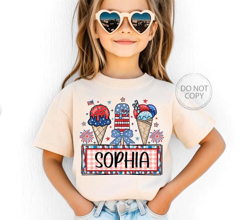May include: A beige t-shirt featuring a patriotic ice cream design with red, white, and blue colors. The design includes ice cream cones, popsicles, and the name "SOPHIA" in a red and white checkered frame. The shirt is worn by a young child.