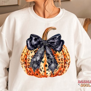 May include: White sweatshirt with a graphic of a pumpkin with a black bow and leopard print. The pumpkin is orange and brown with black lace accents. The text "DESIGN SOUL" is printed on the bottom right corner of the sweatshirt.