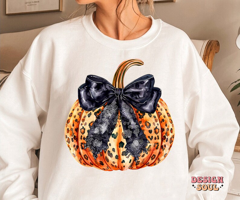 May include: White sweatshirt with a graphic of a pumpkin with a black bow and leopard print. The pumpkin is orange and brown with black lace accents. The text "DESIGN SOUL" is printed on the bottom right corner of the sweatshirt.