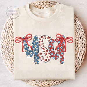 May include: A cream-colored sweatshirt with the word "USA" in red, white, and blue floral fabric. The letters are decorated with red bows.