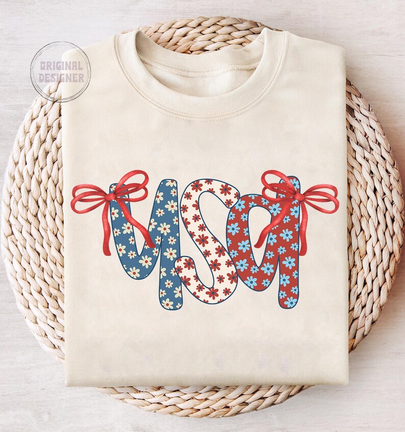 May include: A cream-colored sweatshirt with the word "USA" in red, white, and blue floral fabric. The letters are decorated with red bows.