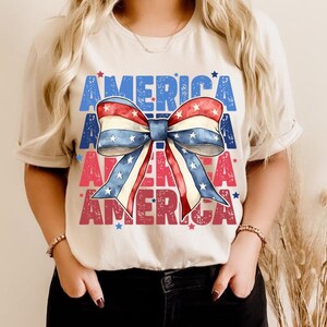 May include: A beige t-shirt featuring a patriotic bow design in red, white, and blue. The word "AMERICA" is printed in blue and red, with stars scattered around the design. A casual and festive top.