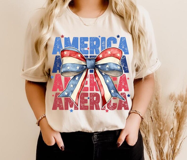 May include: A beige t-shirt featuring a patriotic bow design in red, white, and blue. The word "AMERICA" is printed in blue and red, with stars scattered around the design. A casual and festive top.