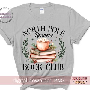 May include: A gray t-shirt with the text "North Pole Readers Book Club" in black. The design features a cup of hot chocolate with marshmallows and a stack of books.