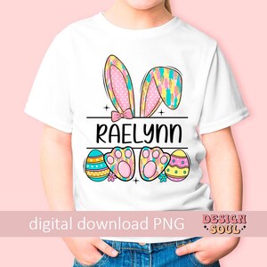 May include: White t-shirt with a pink and yellow bunny ears design, Easter egg accents, and the name "Raelynn" printed on the front.