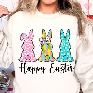 May include: White sweatshirt with a graphic of three Easter bunnies in different patterns. The bunnies are pink, blue, and yellow with white accents. The text "Happy Easter" is printed below the bunnies.