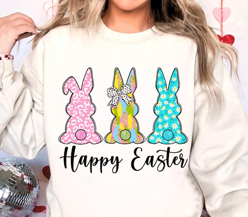 May include: White sweatshirt with a graphic of three Easter bunnies in different patterns. The bunnies are pink, blue, and yellow with white accents. The text "Happy Easter" is printed below the bunnies.