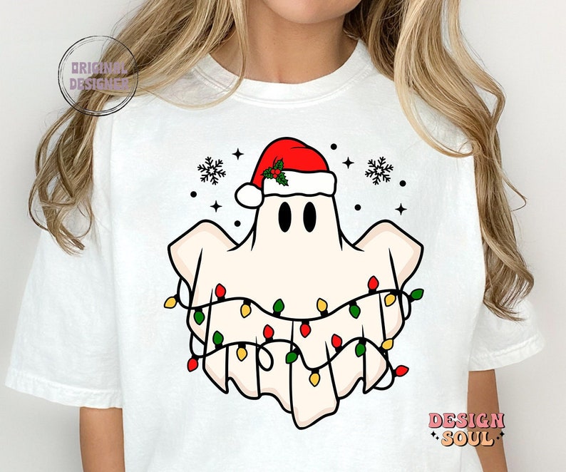 May include: White t-shirt with a cartoon ghost wearing a Santa hat and Christmas lights. The ghost has two black eyes and a red nose.