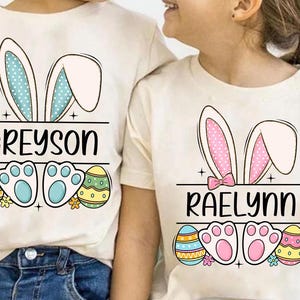 May include: Two white t-shirts with bunny ears and Easter egg designs. The shirts have the names "Greyson" and "Raelynn" printed on them.