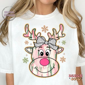 May include: White t-shirt with a pink and green reindeer face graphic. The reindeer has a pink nose, black eyes, and a black and white polka dot bow. The graphic is surrounded by gold glitter and snowflakes.