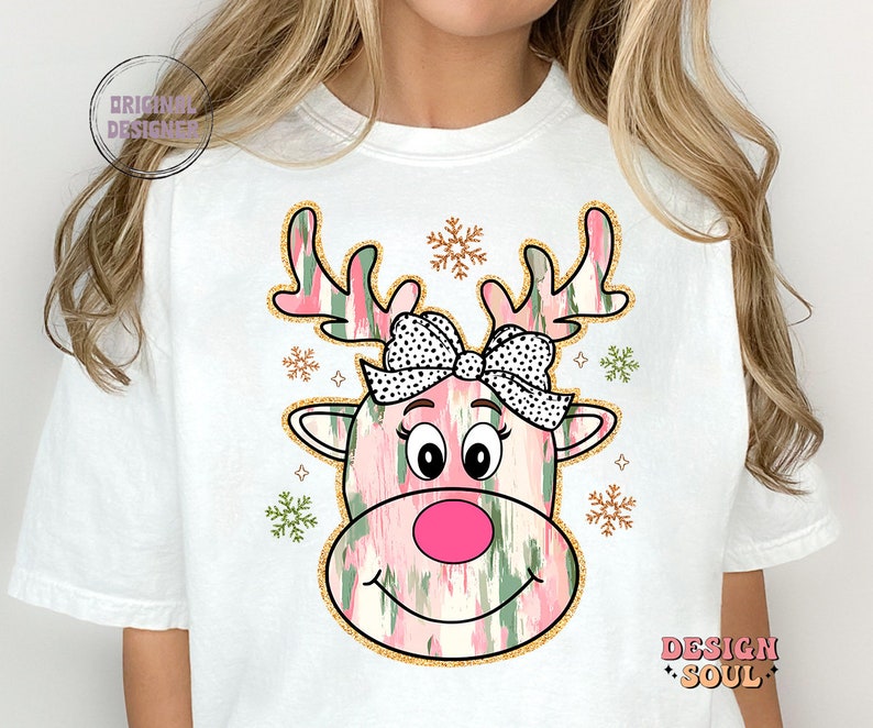 May include: White t-shirt with a pink and green reindeer face graphic. The reindeer has a pink nose, black eyes, and a black and white polka dot bow. The graphic is surrounded by gold glitter and snowflakes.