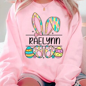 May include: Pink sweatshirt with a bunny ear design, Easter eggs, and the name "RAELynn" printed on the front.