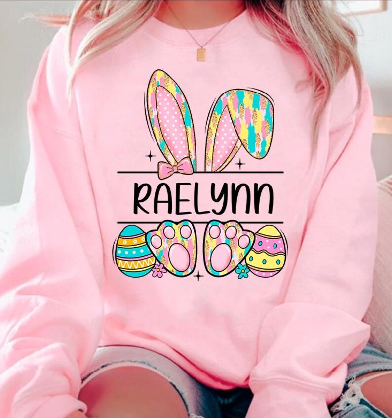 May include: Pink sweatshirt with a bunny ear design, Easter eggs, and the name "RAELynn" printed on the front.
