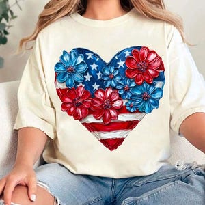 May include: A white t-shirt with a red, white, and blue heart-shaped design. The heart is made up of flowers and an American flag.