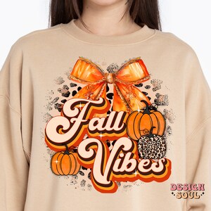 May include: A tan sweatshirt with a retro-style graphic design featuring the text "Fall Vibes" in orange and brown. The design also includes a large orange bow, two pumpkins, and a leopard print pattern.