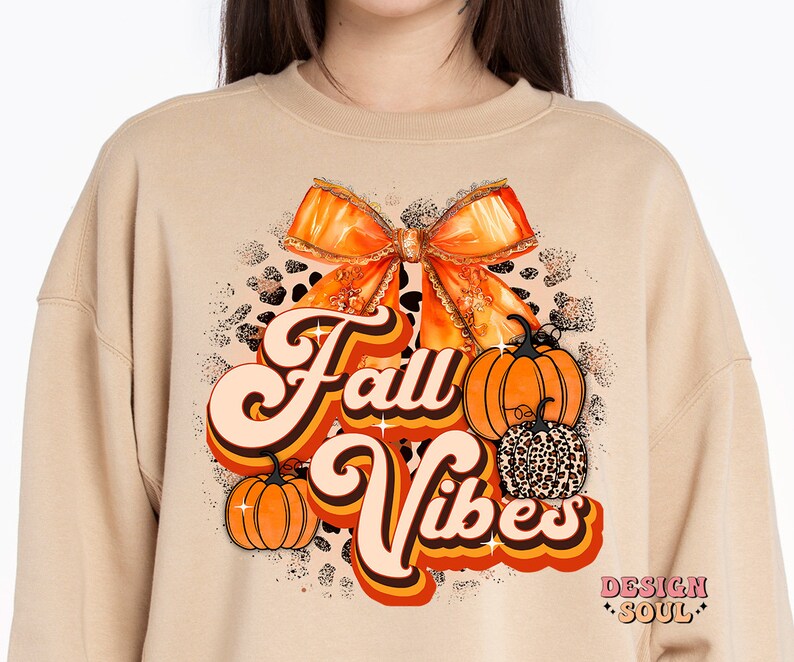 May include: A tan sweatshirt with a retro-style graphic design featuring the text "Fall Vibes" in orange and brown. The design also includes a large orange bow, two pumpkins, and a leopard print pattern.
